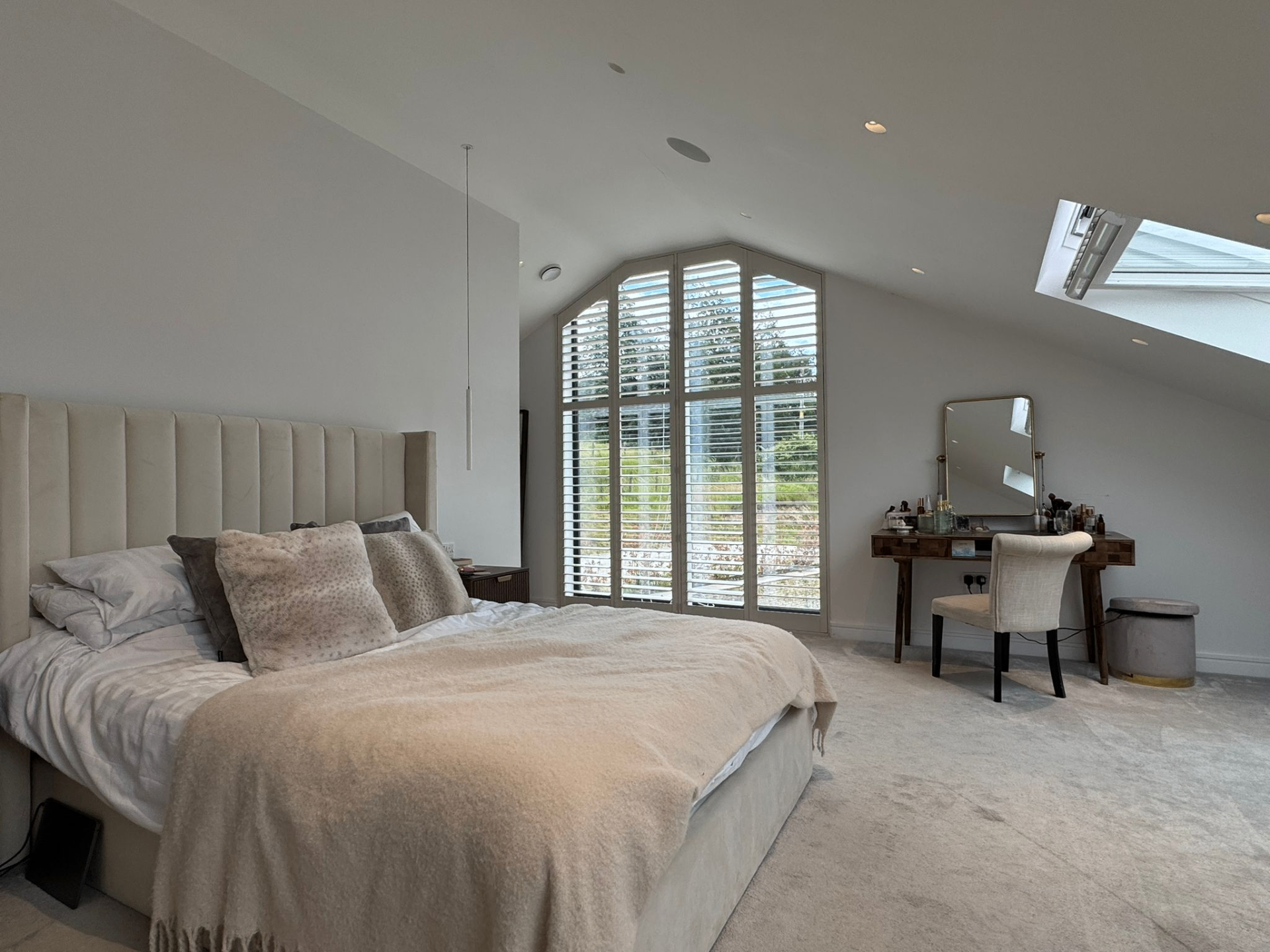 Angled bedroom view showing shaped shutters across a tall pitched window beside a dressing table
