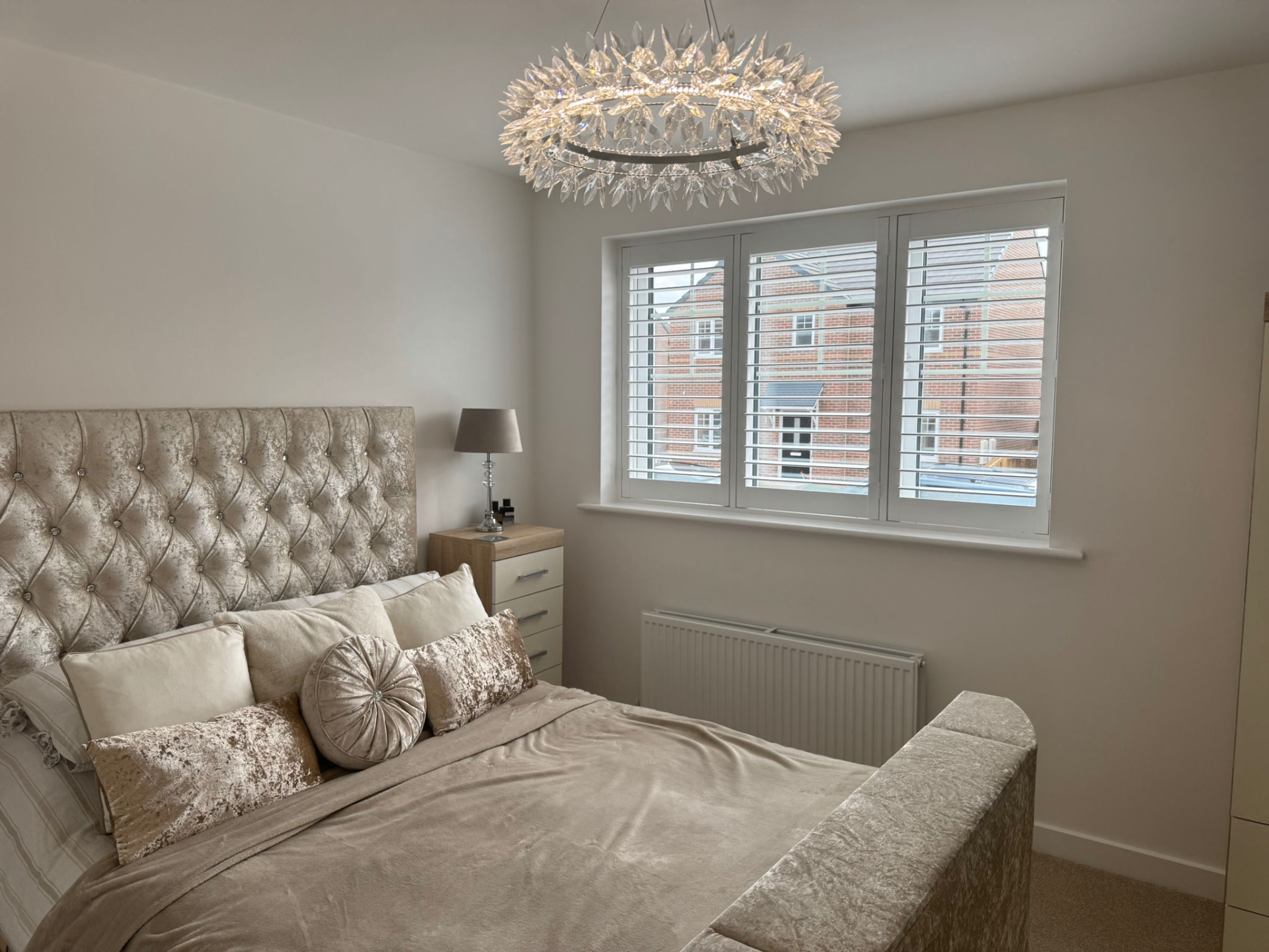 Full height white shutters fitted to a bedroom window with upholstered bed and crystal ceiling light